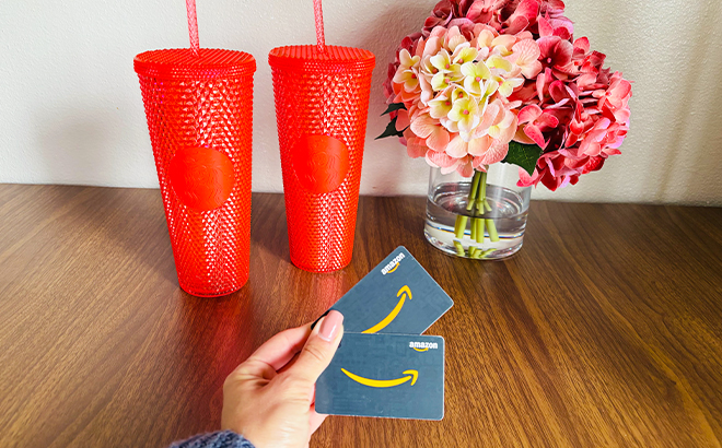 A Person Holding Two Amazon Gift Cards in Front of Two Red Starbucks Tumblers